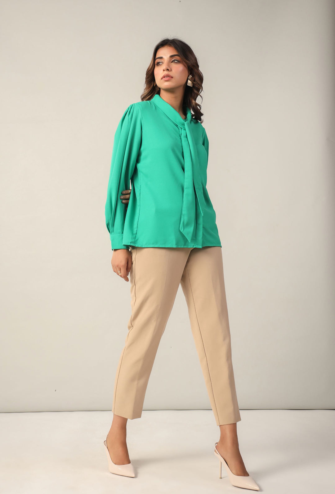 Woman in emerald green tie-neck blouse with puff sleeves, beige trousers, and white heels