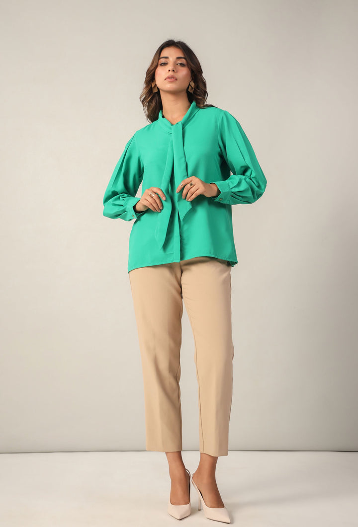 Woman in emerald green tie-neck blouse with long sleeves, paired with beige trousers and heels