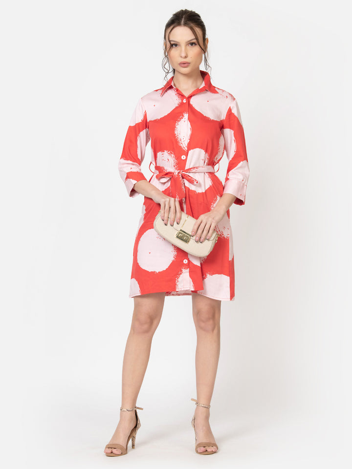 Woman in coral and pink abstract print shirt dress with tie waist, holding clutch, wearing heels