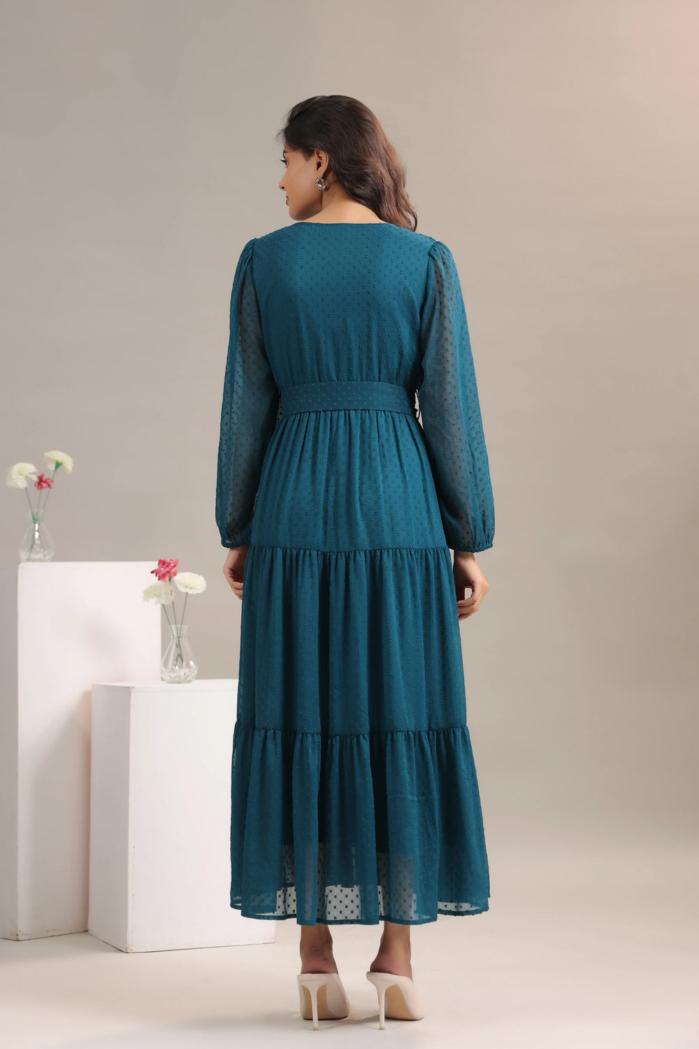 Woman wearing teal tiered maxi dress with sheer long sleeves, standing in studio.