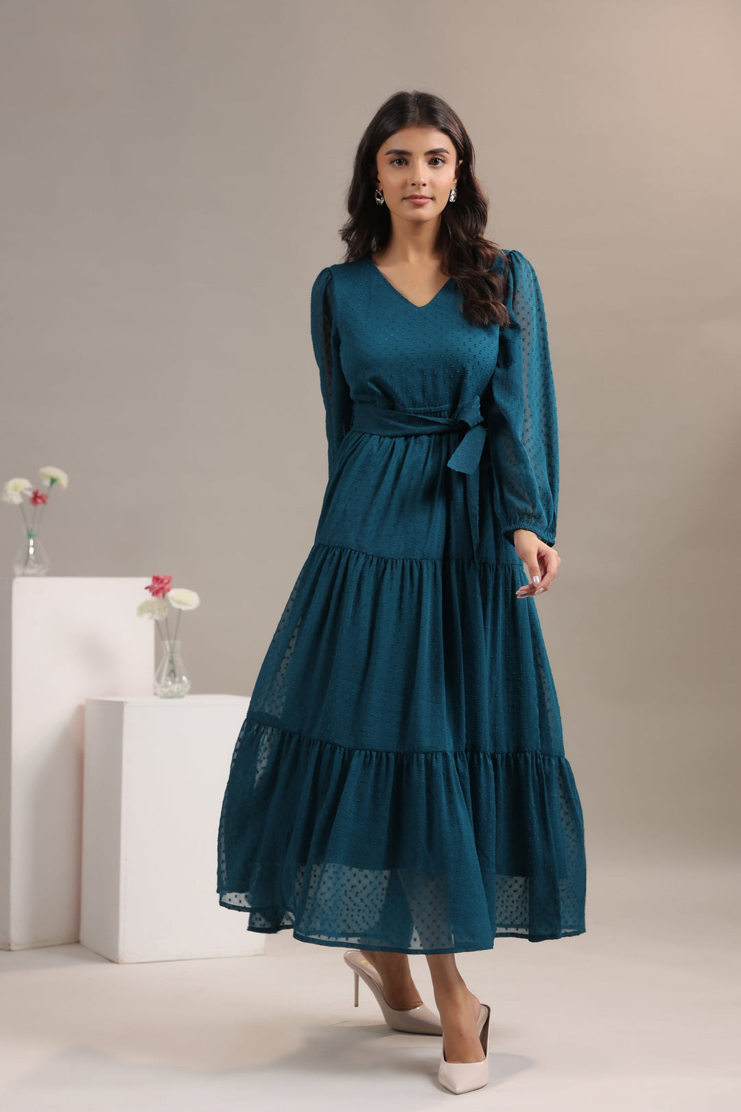 Woman wearing a teal dress with long sleeves and a belt, standing against a neutral background.